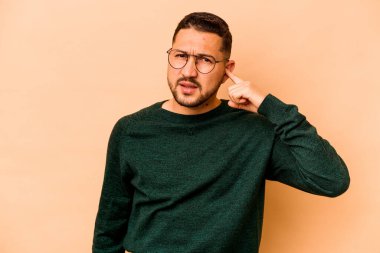 Latin kökenli genç adam bej arka planda izole edilmiş. Kulakları parmaklarıyla kapatıyor. Yüksek sesli bir ortam yüzünden stresli ve çaresiz..