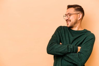 Latin kökenli genç adam bej arka planda izole edilmiş. Kollarını kavuşturarak gülümsüyor..
