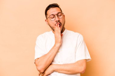 Latin kökenli genç adam bej arka planda izole edilmiş esniyor. Ağzı eliyle kapatan yorgun bir hareket sergiliyor..