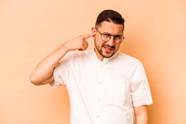 Latin kökenli genç adam bej arka planda izole edilmiş kulakları elleriyle kapatıyor..
