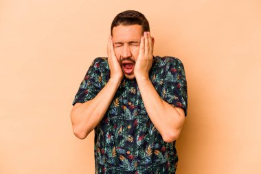 Latin kökenli genç adam bej arka planda izole edilmiş kulakları kulaklarıyla kapatıyor çok fazla ses duymamaya çalışıyor..