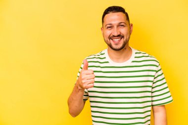 Sarı arka planda izole edilmiş genç İspanyol adam gülümsüyor ve başparmağını kaldırıyor.