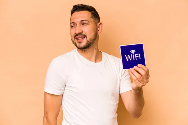 Hispanic man holding wifi placard isolated on beige background looks aside smiling, cheerful and pleasant.