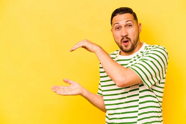 Young hispanic man isolated on yellow background shocked and amazed holding a copy space between hands.