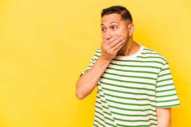 Genç İspanyol adam sarı arka planda izole edilmiş. Düşünceli bir şekilde bir fotokopi odasına bakıyor. Ağzını eliyle kapatıyor..