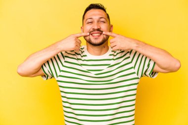 Sarı arka planda izole edilmiş genç İspanyol adam gülümsüyor, parmaklarını ağıza doğrultuyor..