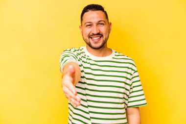 Sarı arka planda izole edilmiş genç İspanyol adam selamlama jesti olarak kameraya elini uzatıyor..