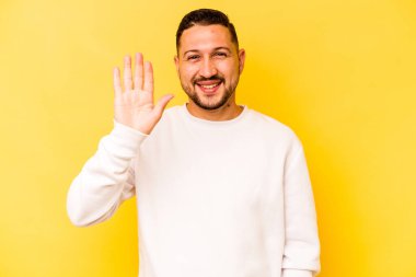 Sarı arka planda izole edilmiş genç İspanyol adam gülümseyerek beş numarayı parmaklarıyla gösteriyor..
