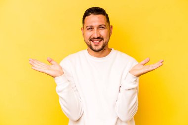 Sarı arka planda izole edilmiş genç İspanyol adam kollarıyla ölçeği yapıyor, mutlu ve kendinden emin hissediyor..