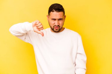 Sarı arka planda izole edilmiş genç İspanyol adam baş parmağını aşağı gösteriyor, hayal kırıklığı konsepti..