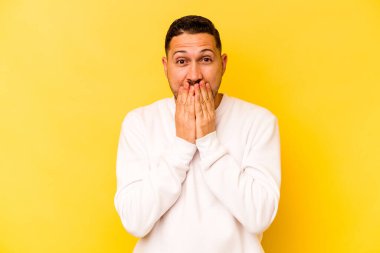 Sarı arka planda izole edilmiş genç İspanyol adam şok olmuş, ağzı ellerle kaplanmış, yeni bir şey keşfetmek için sabırsızlanıyor..