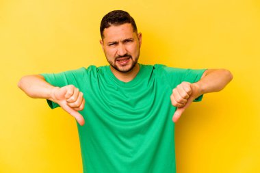 Sarı arka planda izole edilmiş genç İspanyol adam baş parmağını aşağı gösteriyor ve hoşlanmadığını ifade ediyor..