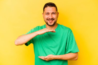 Sarı arka planda izole edilmiş genç İspanyol adam iki eliyle bir şey tutuyor, ürün sunumu..