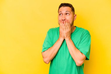 Genç İspanyol adam sarı arka planda izole edilmiş. Düşünceli bir şekilde bir fotokopi odasına bakıyor. Ağzını eliyle kapatıyor..
