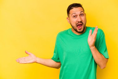 Sarı arka planda izole edilmiş genç İspanyol bir adam avuç içinde fotokopi alanı tutuyor, el üstünde tutuyor. Şaşırmış ve mutlu..