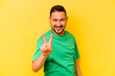 Sarı arka planda izole edilmiş genç İspanyol adam zafer işareti gösteriyor ve gülümsüyor..