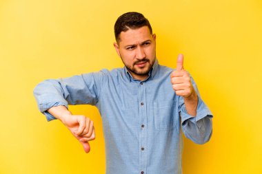Latin kökenli genç adam sarı arka planda izole edilmiş, baş parmağını kaldırıyor, zor bir seçim.