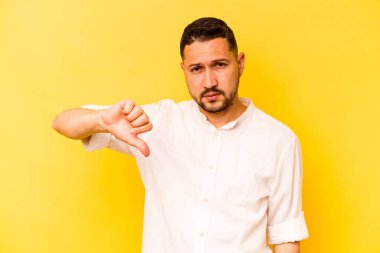 Sarı arka planda izole edilmiş genç İspanyol adam baş parmağını aşağı gösteriyor, hayal kırıklığı konsepti..