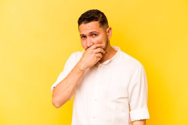 Sarı arka planda izole edilmiş genç İspanyol adam elleri endişeli bir şekilde ağzı örtüyor..