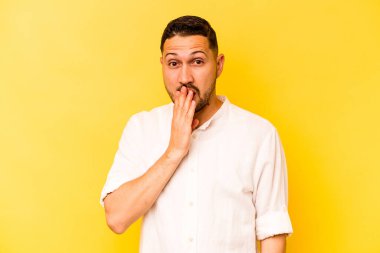 Sarı arka planda izole edilmiş genç İspanyol adam şok olmuş, ağzı ellerle kaplanmış, yeni bir şey keşfetmek için sabırsızlanıyor..