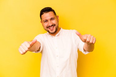 Genç İspanyol adam sarı arka planda izole edilmiş. Baş parmağını kaldırıyor, gülümsüyor ve kendinden emin..