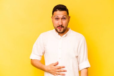 Sarı arka planda izole edilmiş genç İspanyol adam karnına dokunuyor, nazikçe gülümsüyor, yemek yiyor ve tatmin oluyor..