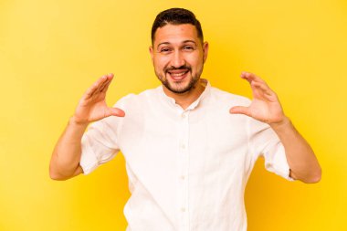 İspanyol asıllı genç adam sarı arka planda izole edilmiş. Elinde avuç içi olan bir şeyle kameraya sunuyor..