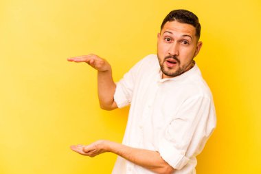 Sarı arka planda izole edilmiş genç İspanyol adam iki eliyle bir şey tutuyor, ürün sunumu..