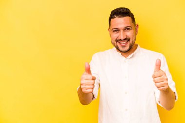 Sarı arka planda izole edilmiş genç İspanyol adam gülümsüyor ve başparmağını kaldırıyor.