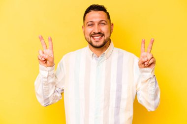 Sarı arka planda izole edilmiş genç İspanyol adam zafer işareti gösteriyor ve gülümsüyor..