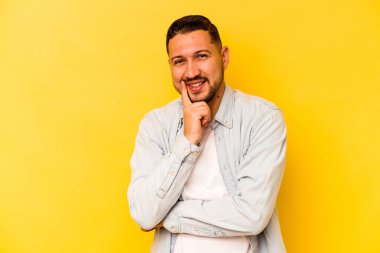 Sarı arka planda izole olmuş genç İspanyol adam gülümsüyor ve kendinden emin, elleriyle çeneye dokunuyor..