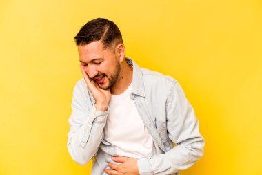 Sarı arka planda izole edilmiş genç İspanyol adam mutlu bir şekilde gülüyor ve ellerini karnının üstünde tutarak eğleniyor..