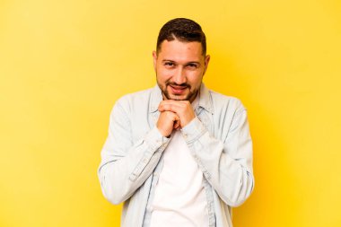 Sarı arka planda izole edilmiş genç İspanyol adam ellerini çenesinin altında tutuyor ve mutlu bir şekilde yan tarafa bakıyor..