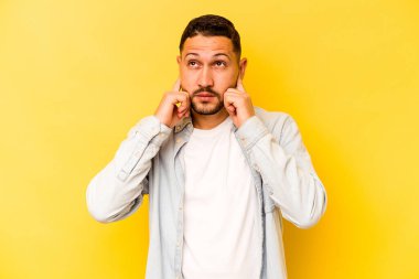 Sarı arka planda izole edilmiş, parmaklarıyla kulaklarını kapatan, stresli ve çaresiz genç bir İspanyol..