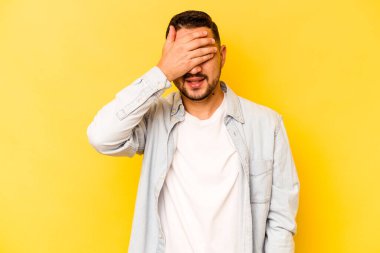 Sarı arka planda izole edilmiş genç İspanyol adam elleriyle gözlerini kapatıyor, geniş bir gülümsemeyle bir sürpriz bekliyor..
