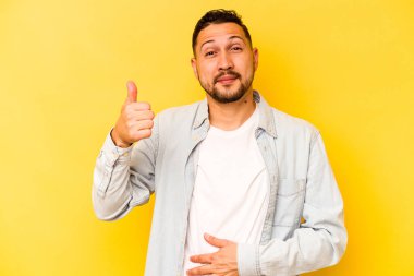 Sarı arka planda izole edilmiş genç İspanyol adam karnına dokunuyor, nazikçe gülümsüyor, yemek yiyor ve tatmin oluyor..