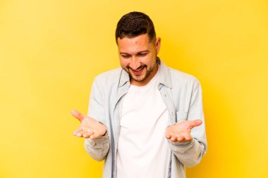 İspanyol asıllı genç adam sarı arka planda izole edilmiş. Elinde avuç içi olan bir şeyle kameraya sunuyor..