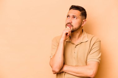 Latin kökenli genç adam bej arka planda izole edilmiş şüpheli ve şüpheci bir ifadeyle yana bakıyor..