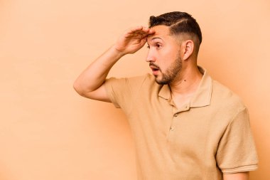 Latin kökenli genç adam bej arka planda izole edilmiş, uzaklara bakıyor ve elini alnına koyuyor..