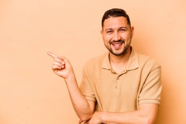 Latin kökenli genç adam, bej arka planda izole edilmiş. Gülümsüyor, işaret parmağıyla işaret ediyor..