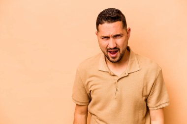 Latin kökenli genç adam bej arka planda izole edilmiş çok kızgın, öfkeli, hayal kırıklığına uğramış bağırıyor..