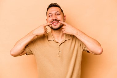 Latin kökenli genç adam bej arka planda izole edilmiş gülümsüyor, parmaklarını ağıza doğrultuyor..