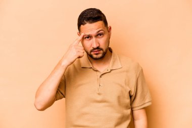 Latin kökenli genç bir adam bej arka planda izole edilmiş. Şakağı parmağıyla işaret ediyor, düşünüyor ve bir göreve odaklanıyor..