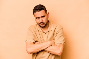 Latin kökenli genç adam bej arka planda izole edilmiş hoşnutsuzca kaşlarını çatmış, kollarını kavuşturmuş..