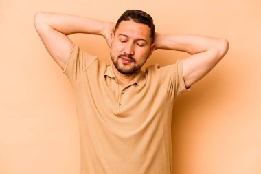 Latin kökenli genç adam bej arka planda izole edilmiş. Kendine güveniyor, elleri başının arkasında..