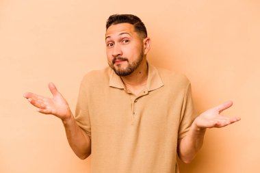 Latin kökenli genç bir adam, bej arka planda izole edilmiş sorgulama jestinde şüphe ediyor ve omuz silkiyor..