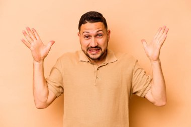 Latin kökenli genç adam bej arka planda izole edilmiş gökyüzüne bağırıyor, yukarı bakıyor, hayal kırıklığına uğramış..