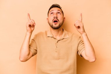Latin kökenli genç adam bej arka planda izole edilmiş. Ağzı açık, üst tarafı işaret ediyor..