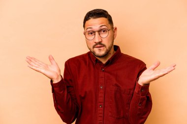 Latin kökenli genç adam bej arka planda izole edilmiş. Kopya alanını tutmak için kafası karışık ve omuz silkiyor..