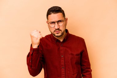 Latin kökenli genç adam bej arka planda izole edilmiş. Kameraya yumruk, agresif yüz ifadesi gösteriyor..